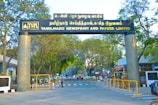 An entrance gate of a company with the name Tamilnadu Newsprint and Papers Limited displayed prominently. The gate is flanked by two large columns and surrounded by trees. People are seen walking near the entrance, and security personnel are present. There are vehicles parked or moving around, indicating an active setting.