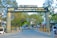 An entrance gate of a company with the name Tamilnadu Newsprint and Papers Limited displayed prominently. The gate is flanked by two large columns and surrounded by trees. People are seen walking near the entrance, and security personnel are present. There are vehicles parked or moving around, indicating an active setting.