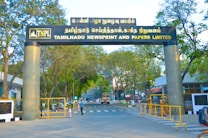 An entrance gate of a company with the name Tamilnadu Newsprint and Papers Limited displayed prominently. The gate is flanked by two large columns and surrounded by trees. People are seen walking near the entrance, and security personnel are present. There are vehicles parked or moving around, indicating an active setting.