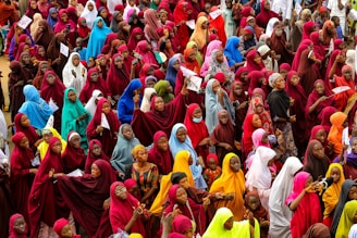 A large crowd of women dressed in brightly colored hijabs gather closely together. The hijabs are in various shades including red, blue, green, yellow, and orange. Some women are holding papers, and others appear to be interacting with each other.