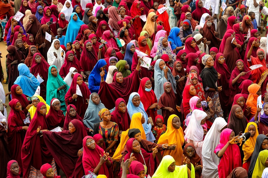 A large crowd of women dressed in brightly colored hijabs gather closely together. The hijabs are in various shades including red, blue, green, yellow, and orange. Some women are holding papers, and others appear to be interacting with each other.