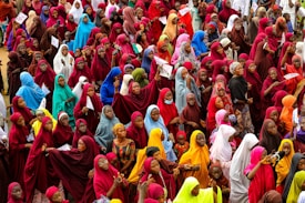 A large crowd of women dressed in brightly colored hijabs gather closely together. The hijabs are in various shades including red, blue, green, yellow, and orange. Some women are holding papers, and others appear to be interacting with each other.
