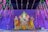 A colorful statue of the Hindu deity Ganesha is adorned with intricate garments and garlands of fresh flowers. The backdrop features a vibrant purple curtain and hanging floral decorations, creating a festive atmosphere. Two oil lamps are placed symmetrically on each side of the statue.