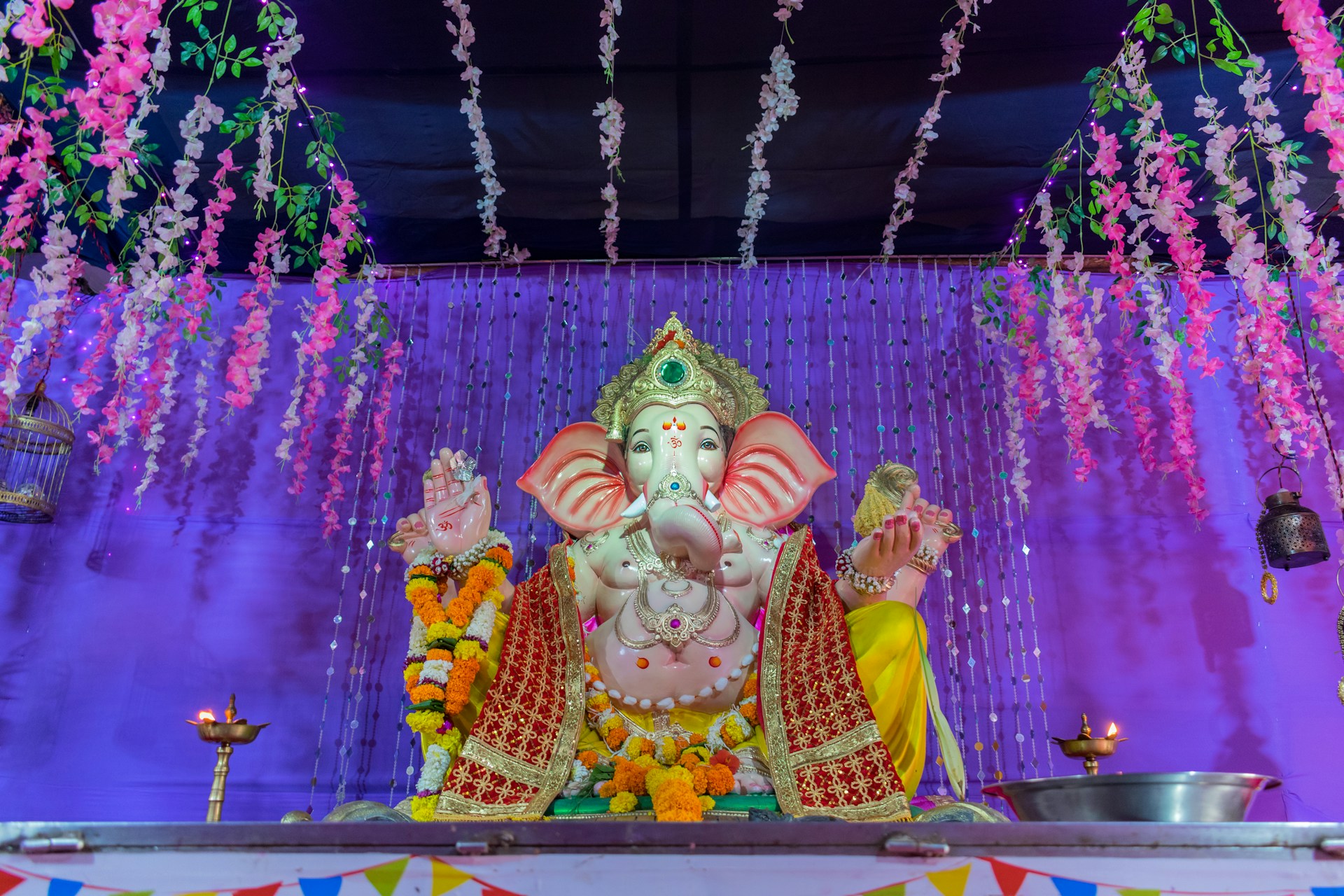 A colorful statue of the Hindu deity Ganesha is adorned with intricate garments and garlands of fresh flowers. The backdrop features a vibrant purple curtain and hanging floral decorations, creating a festive atmosphere. Two oil lamps are placed symmetrically on each side of the statue.