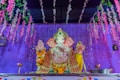 A colorful statue of the Hindu deity Ganesha is adorned with intricate garments and garlands of fresh flowers. The backdrop features a vibrant purple curtain and hanging floral decorations, creating a festive atmosphere. Two oil lamps are placed symmetrically on each side of the statue.