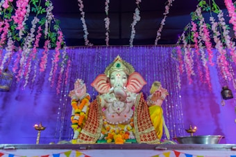 A colorful statue of the Hindu deity Ganesha is adorned with intricate garments and garlands of fresh flowers. The backdrop features a vibrant purple curtain and hanging floral decorations, creating a festive atmosphere. Two oil lamps are placed symmetrically on each side of the statue.