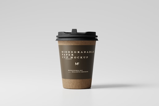 A paper coffee cup with a black lid stands upright against a plain, light gray background. The cup is labeled as a biodegradable paper cup mockup with elegant white text printed on an olive green band around its upper section. The bottom part of the cup shows a natural brown color, typical of kraft paper.