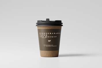A paper coffee cup with a black lid stands upright against a plain, light gray background. The cup is labeled as a biodegradable paper cup mockup with elegant white text printed on an olive green band around its upper section. The bottom part of the cup shows a natural brown color, typical of kraft paper.
