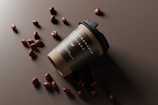 A biodegradable coffee cup with a black lid is tilted on its side on a dark surface. Scattered around the cup are several coffee beans that create a visually appealing arrangement.