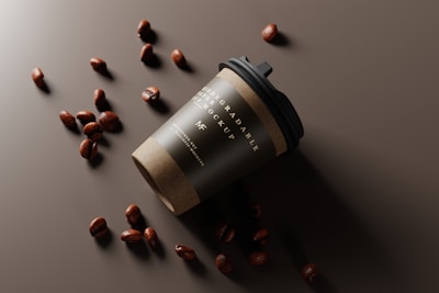 A biodegradable coffee cup with a black lid is tilted on its side on a dark surface. Scattered around the cup are several coffee beans that create a visually appealing arrangement.