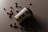 A biodegradable coffee cup with a black lid is tilted on its side on a dark surface. Scattered around the cup are several coffee beans that create a visually appealing arrangement.