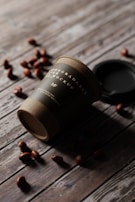 A biodegradable paper cup lies on its side on a rustic wooden surface, surrounded by scattered coffee beans. The cup has a dark label with white text and a partially visible logo. A black lid is placed near the cup.