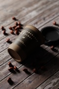 A biodegradable paper cup lies on its side on a rustic wooden surface, surrounded by scattered coffee beans. The cup has a dark label with white text and a partially visible logo. A black lid is placed near the cup.