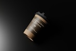 A disposable paper coffee cup with a black lid rests against a dark background. The cup has an earthy brown band with white text indicating it is a biodegradable paper cup mockup. The lighting casts a smooth gradient, creating shadows and highlights on the cup, giving it a realistic appearance.