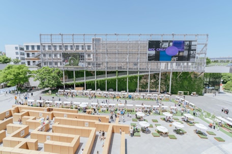 An architectural structure forms the backdrop of an outdoor event featuring a maze constructed from cardboard. The scene includes people walking around and interacting, with various booths or stalls set up along the perimeter. Lush greenery and trees add natural elements, while a large screen displays digital content. Tables with umbrellas and seating arrangements are scattered across the foreground, providing areas for relaxation.