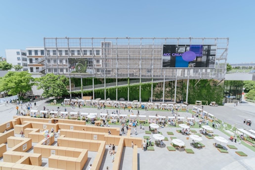 An architectural structure forms the backdrop of an outdoor event featuring a maze constructed from cardboard. The scene includes people walking around and interacting, with various booths or stalls set up along the perimeter. Lush greenery and trees add natural elements, while a large screen displays digital content. Tables with umbrellas and seating arrangements are scattered across the foreground, providing areas for relaxation.