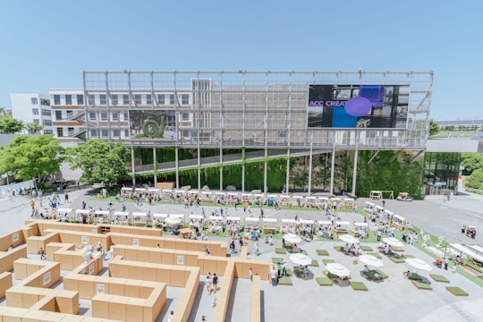 An architectural structure forms the backdrop of an outdoor event featuring a maze constructed from cardboard. The scene includes people walking around and interacting, with various booths or stalls set up along the perimeter. Lush greenery and trees add natural elements, while a large screen displays digital content. Tables with umbrellas and seating arrangements are scattered across the foreground, providing areas for relaxation.