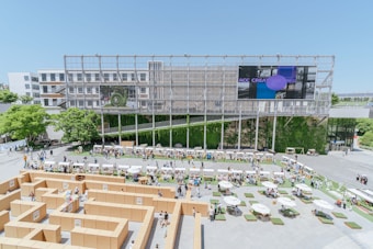 An architectural structure forms the backdrop of an outdoor event featuring a maze constructed from cardboard. The scene includes people walking around and interacting, with various booths or stalls set up along the perimeter. Lush greenery and trees add natural elements, while a large screen displays digital content. Tables with umbrellas and seating arrangements are scattered across the foreground, providing areas for relaxation.