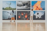 A modern art gallery features six large, surreal photographs on the wall. Each image presents intriguing and dreamlike scenes, including an object floating above a hill, a person engulfed in flames, and abstract shapes. Two visitors, one in a dress and another in casual wear, observe the artwork while standing on a polished wooden floor.