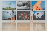 A modern art gallery features six large, surreal photographs on the wall. Each image presents intriguing and dreamlike scenes, including an object floating above a hill, a person engulfed in flames, and abstract shapes. Two visitors, one in a dress and another in casual wear, observe the artwork while standing on a polished wooden floor.