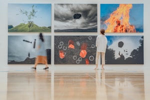A modern art gallery features six large, surreal photographs on the wall. Each image presents intriguing and dreamlike scenes, including an object floating above a hill, a person engulfed in flames, and abstract shapes. Two visitors, one in a dress and another in casual wear, observe the artwork while standing on a polished wooden floor.