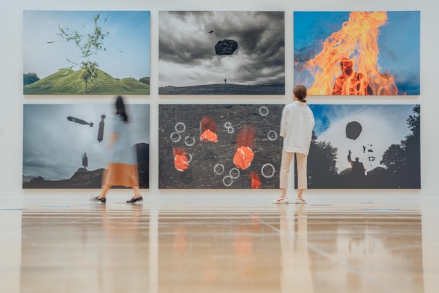 A modern art gallery features six large, surreal photographs on the wall. Each image presents intriguing and dreamlike scenes, including an object floating above a hill, a person engulfed in flames, and abstract shapes. Two visitors, one in a dress and another in casual wear, observe the artwork while standing on a polished wooden floor.