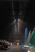 A spacious auditorium features a stage on the right with multiple spotlights shining in different colors, including blue and green. In the audience seating area on the left, several people watch attentively. A speaker or performer stands at a microphone stand in the center of the stage. The ceiling is high and equipped with numerous rigging structures and lighting equipment.