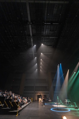 A spacious auditorium features a stage on the right with multiple spotlights shining in different colors, including blue and green. In the audience seating area on the left, several people watch attentively. A speaker or performer stands at a microphone stand in the center of the stage. The ceiling is high and equipped with numerous rigging structures and lighting equipment.