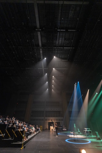 A spacious auditorium features a stage on the right with multiple spotlights shining in different colors, including blue and green. In the audience seating area on the left, several people watch attentively. A speaker or performer stands at a microphone stand in the center of the stage. The ceiling is high and equipped with numerous rigging structures and lighting equipment.