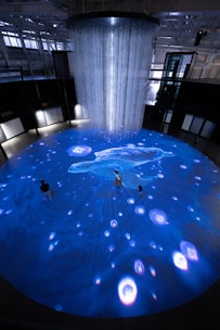 An immersive digital art installation features an illuminated floor with aquatic imagery, including a large digital representation of a marine animal. Several people are interacting with the space, walking on the projection. Above, a cascade of lights or thin strands descends from the ceiling, creating a sense of depth and fluidity in the ambient lighting.