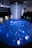 An immersive digital art installation features an illuminated floor with aquatic imagery, including a large digital representation of a marine animal. Several people are interacting with the space, walking on the projection. Above, a cascade of lights or thin strands descends from the ceiling, creating a sense of depth and fluidity in the ambient lighting.