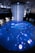 An immersive digital art installation features an illuminated floor with aquatic imagery, including a large digital representation of a marine animal. Several people are interacting with the space, walking on the projection. Above, a cascade of lights or thin strands descends from the ceiling, creating a sense of depth and fluidity in the ambient lighting.