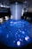 An immersive digital art installation features an illuminated floor with aquatic imagery, including a large digital representation of a marine animal. Several people are interacting with the space, walking on the projection. Above, a cascade of lights or thin strands descends from the ceiling, creating a sense of depth and fluidity in the ambient lighting.