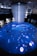 An immersive digital art installation features an illuminated floor with aquatic imagery, including a large digital representation of a marine animal. Several people are interacting with the space, walking on the projection. Above, a cascade of lights or thin strands descends from the ceiling, creating a sense of depth and fluidity in the ambient lighting.