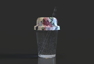 A transparent cup filled with a clear liquid, possibly sparkling water, featuring noticeable bubbles. The cup has a lid with a colorful, abstract pattern consisting of various shapes and colors, and a straw emerges from the top.