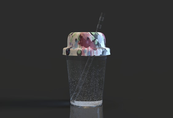 A transparent cup filled with a clear liquid, possibly sparkling water, featuring noticeable bubbles. The cup has a lid with a colorful, abstract pattern consisting of various shapes and colors, and a straw emerges from the top.