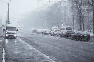 A city street covered in light snowfall, with several cars and vans driving along. The road is wet and slushy, and the air appears dense with falling snowflakes. Streetlights and traffic signals are visible, as well as bare trees lining the sidewalks.