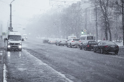 A city street covered in light snowfall, with several cars and vans driving along. The road is wet and slushy, and the air appears dense with falling snowflakes. Streetlights and traffic signals are visible, as well as bare trees lining the sidewalks.