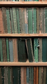 A shelf filled with a collection of worn and aged books, most of which have green and beige spines. The covers are tattered and frayed, suggesting significant age and use. The titles are printed in various languages, and the books are arranged vertically.
