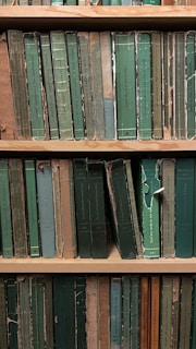 A shelf filled with a collection of worn and aged books, most of which have green and beige spines. The covers are tattered and frayed, suggesting significant age and use. The titles are printed in various languages, and the books are arranged vertically.