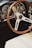 A classic car interior featuring a wooden steering wheel and a dashboard filled with vintage gauges and dials. The seating is upholstered in light-colored leather.