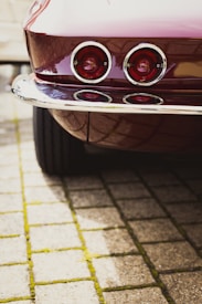A vintage car's rear view is shown, featuring two round tail lights with chrome surrounds. The vehicle's glossy maroon paint reflects light, and the scene includes paving stones with moss growing between them, adding texture to the foreground.