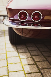 A vintage car's rear view is shown, featuring two round tail lights with chrome surrounds. The vehicle's glossy maroon paint reflects light, and the scene includes paving stones with moss growing between them, adding texture to the foreground.