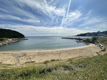 A tranquil beach scene with a sandy shore gently meeting a calm, blue sea. The horizon displays a partly cloudy sky with a radiant beam of sunlight streaming down, creating a serene atmosphere. To the sides, rocky edges and lush green hills frame the cove, accompanied by sparse buildings in the distance.