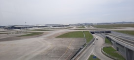 An expansive view of an airport runway with several airplanes positioned near terminal buildings in the distance. The foreground features wide runways and taxiways with clear markings, and there is a road running parallel with a few vehicles moving along it. The sky is clear with minimal cloud cover, creating a bright atmosphere.