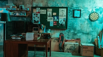 A dimly lit room with a vintage aesthetic, featuring an old wooden desk cluttered with papers, a box, and a retro computer monitor. The background showcases a bulletin board with various sketches and photographs attached. Nearby, shelves contain old books and electronic equipment. A dartboard hangs on the wall, adding to the retro feel.