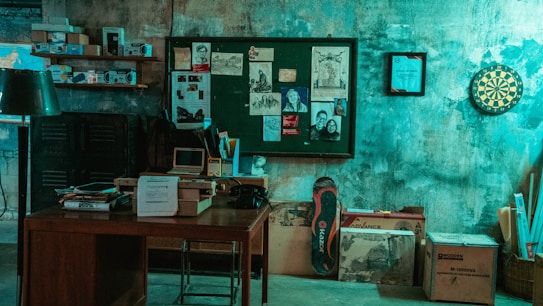 A dimly lit room with a vintage aesthetic, featuring an old wooden desk cluttered with papers, a box, and a retro computer monitor. The background showcases a bulletin board with various sketches and photographs attached. Nearby, shelves contain old books and electronic equipment. A dartboard hangs on the wall, adding to the retro feel.