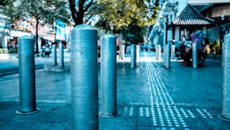 A row of metallic bollards lines a city street, leading towards a busy urban scene featuring pedestrians and trees. The surface of the bollards has a textured pattern, and a tactile guiding pathway runs parallel, indicating accessibility features. Buildings and a small pavilion with a tiled roof are visible in the background, contributing to the bustling yet orderly environment.