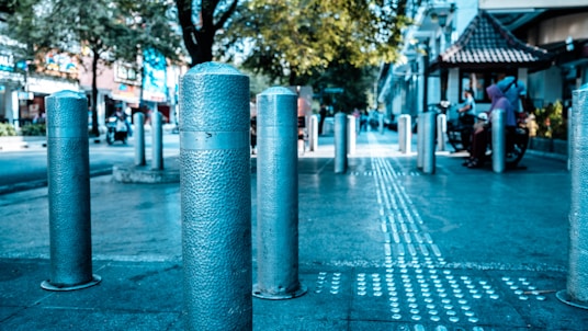 A row of metallic bollards lines a city street, leading towards a busy urban scene featuring pedestrians and trees. The surface of the bollards has a textured pattern, and a tactile guiding pathway runs parallel, indicating accessibility features. Buildings and a small pavilion with a tiled roof are visible in the background, contributing to the bustling yet orderly environment.