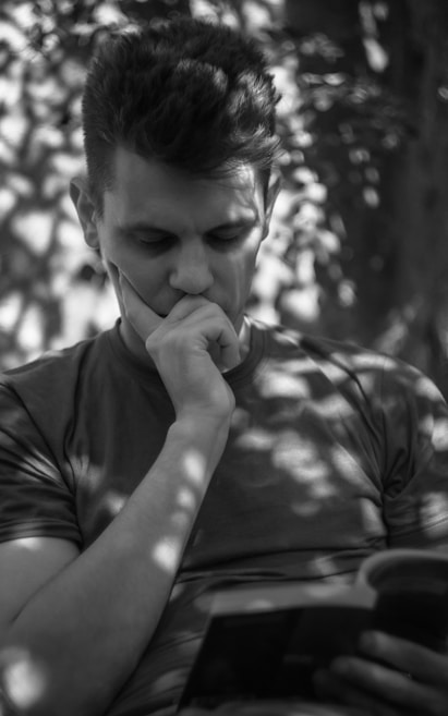 A person is sitting and appears to be deeply absorbed in thought while looking at a book. The lighting creates an interesting pattern of light and shadow on their face and clothing, suggesting they are outdoors under a tree.